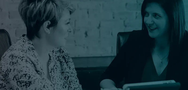 A desaturated image with a left to right gradient from grey to blue. Two women sitting a desk looking at each other while holding a book or tablet.