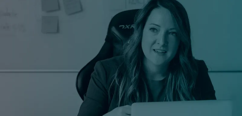 A desaturated image with a left to right gradient from grey to blue. A woman sitting in a chair looking at the camera with a laptop on the desk and a collection of coloured sticky notes in the background.