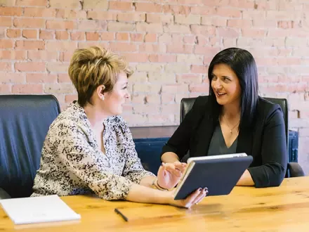 Two women sitting a desk looking at each other while holding a book or tablet.