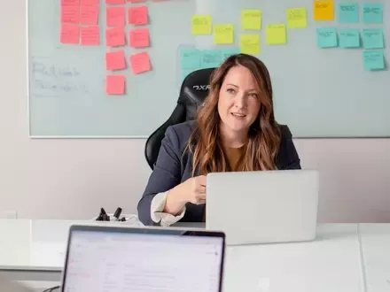 A woman sitting in a chair looking at the camera with a laptop on the desk and a collection of coloured sticky notes in the background.