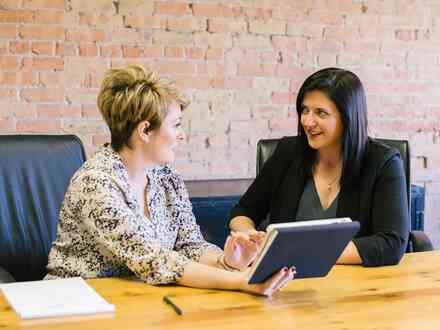 Two women sitting a desk looking at each other while holding a book or tablet