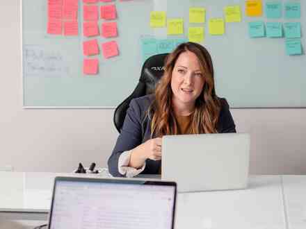 A woman sitting in a chair looking at the camera with a laptop on the desk and a collection of coloured sticky notes in the background.